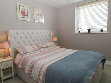 A bedroom with a bed with striped bedding and a blue blanket two bedside tables with lamps and a window with blinds and plants on the windowsill at 7 Warren Edge Close in Bournemouth