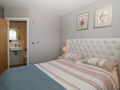 A bedroom with a bed with striped bedding and pillows a lamp on a bedside table two framed floral pictures on the wall and an open door leading to a bathroom at 7 Warren Edge Close Bournemouth