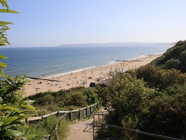 A sandy beach by the sea with people sunbathing and walking with greenery and stairs in the foreground at 7 Warren Edge Close in Bournemouth