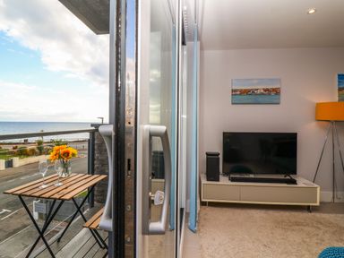 A balcony with a wooden table and two glasses overlooking the sea and a living room with a TV on a stand and a floor lamp at 7 Warren Edge Close in Bournemouth
