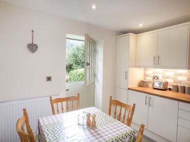 A kitchen with wooden chairs around a table covered with a tablecloth and white cabinets with wooden countertop at Beaver Grove Cottage in Betws-Y-Coed