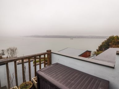 A balcony with a wooden railing overlooking a body of water with hills and buildings in the distance at Cheriton in Pembroke Dock