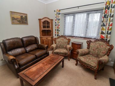 A living room with a brown leather sofa two floral armchairs a wooden coffee table a wooden cabinet a small wooden chest of drawers and a window with lace curtains and patterned drapes at Cheriton in Pembroke Dock