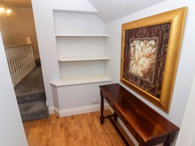 A hallway with wooden floor a wooden table a framed floral painting wall shelves and carpeted stairs at Cheriton Pembroke Dock