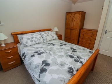 A bedroom with a double bed with patterned bedding two bedside tables with lamps and wooden wardrobe and chest of drawers at Cheriton in Pembroke Dock