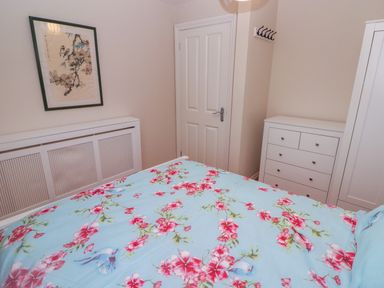 A bedroom with a bed with floral bedding a white dresser a white wardrobe a door and a framed artwork on the wall at Cheriton in Pembroke Dock