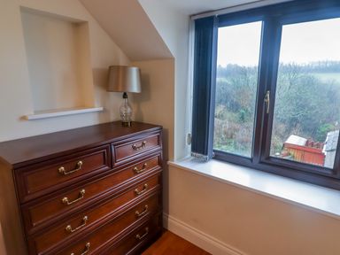 A room corner with a wooden chest of drawers and a lamp next to a window showing an outdoor view at Cheriton in Pembroke Dock