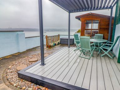An outdoor patio area with a table and chairs under a pergola and a wooden shed near a body of water at Cheriton in Pembroke Dock