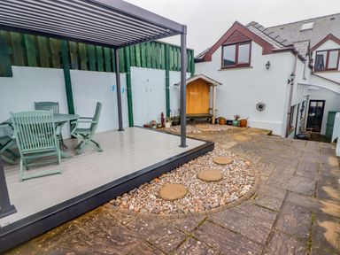 An outdoor patio area with green wooden chairs and table under a pergola near a white house with potted plants and stepping stones at Cheriton in Pembroke Dock