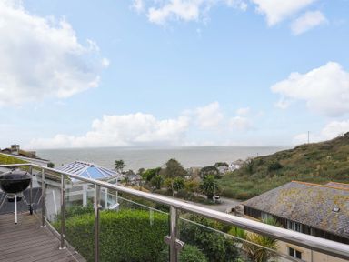 A balcony with glass railing and a barbecue overlooking houses hills and the sea at Waves Edge in Looe