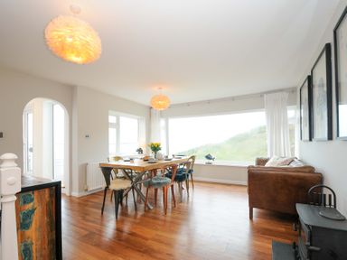 A dining room with wooden table and chairs next to a large window and a brown sofa at Waves Edge in Looe