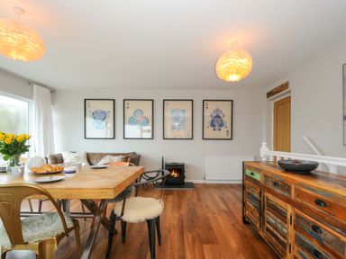 A dining room with a wooden table, chairs, a wooden sideboard, a sofa, framed pictures on the wall, and a small fireplace at Waves Edge in Looe