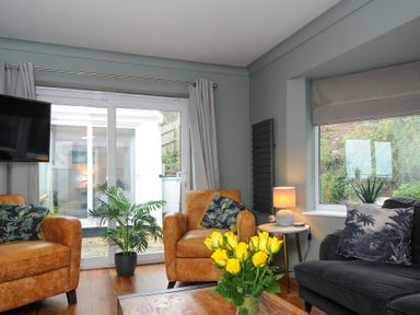 A living room with two brown armchairs a dark gray sofa yellow flowers on a table and sliding glass doors at Waves Edge in Looe