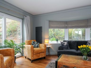 A living room with armchairs a sofa coffee table and windows overlooking plants at Waves Edge in Looe
