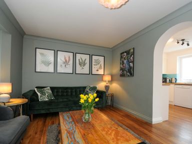 A living room with a green sofa, framed botanical prints on the wall, a wooden coffee table with yellow flowers, and a view into a kitchen at Waves Edge in Looe