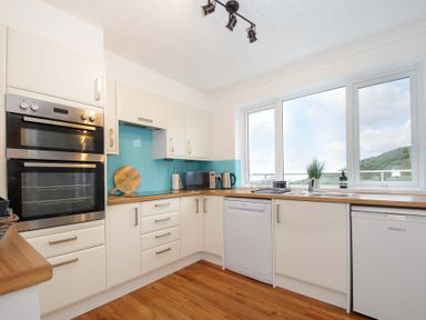 A kitchen with white cabinets wooden countertops appliances and a window showing a hillside at Waves Edge in Looe