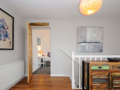 A hallway with wooden flooring a partially open door showing a bedroom a wooden sideboard a white railing and two paintings at Waves Edge in Looe