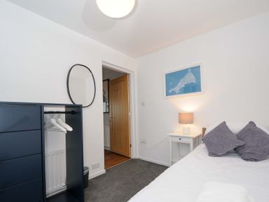 A bedroom with a bed, bedside table with a lamp, blue map picture on the wall, wardrobe with hangers, and an open wooden door at Waves Edge in Looe