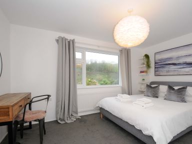 A bedroom with a gray bed and white bedding next to a window with gray curtains and a wooden desk with a chair at Waves Edge in Looe