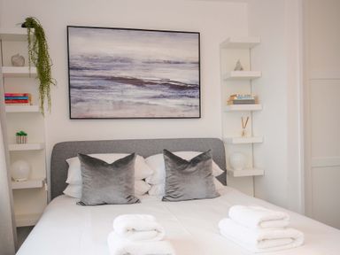A bedroom with a bed with gray pillows white towels on the bed and shelves with books and decorations at Waves Edge in Looe