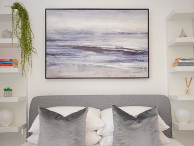 A bed with gray pillows and white sheets below a large abstract painting with blue and purple hues at Waves Edge in Looe