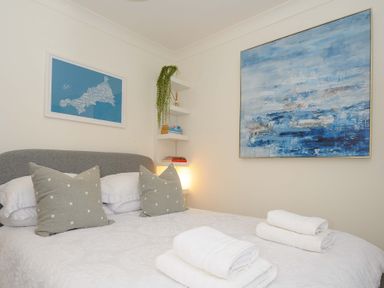 A bedroom with a bed with white bedding and pillows a blue abstract painting on the wall and shelves with books and a plant at Waves Edge in Looe