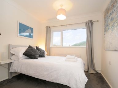 A bedroom with a double bed with white bedding and black pillows next to a window with grey curtains at Waves Edge in Looe