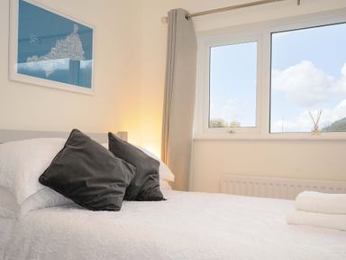 A bedroom with a bed with white bedding and two dark pillows a window with curtains and a framed picture on the wall at Waves Edge in Looe