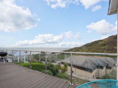 A balcony with glass railing overlooking houses trees a hill and the sea at Waves Edge in Looe