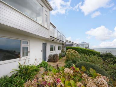 An exterior view of a white house with large windows a garden path and shrubs by the sea at Waves Edge in Looe