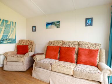 A living room with a floral sofa and armchair with red cushions and paintings on the wall at Fox Cove near St Merryn