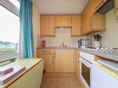 A kitchen with wooden cabinets a sink a stove a microwave and a window with blue curtains at Fox Cove near St Merryn