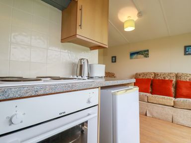 A kitchen with an electric stove toaster kettle and refrigerator next to a living room with a sofa and orange cushions at Fox Cove near St Merryn