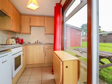 A kitchen with wooden cabinets a window with red curtains and a foldable wooden table near a picnic bench outside at Pepper Cove near St Merryn