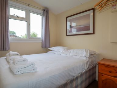 A bedroom with a bed made with white linens towels on the bed a wooden nightstand a window with curtains and a framed picture on the wall at Pepper Cove near St Merryn