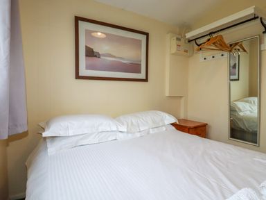 A bedroom with a double bed white bedding a framed picture above the bed a small wooden bedside table hangers on a rod and a wall mirror at Pepper Cove near St Merryn