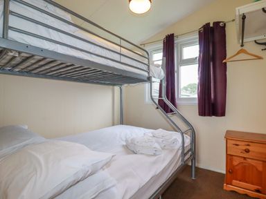 A bedroom with a metal bunk bed a wooden nightstand and a window with curtains at Pepper Cove near St Merryn