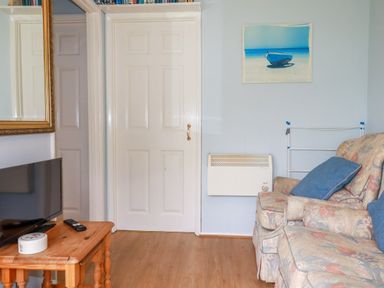 A room with a floral patterned sofa with blue cushions a wooden side table with a TV and remote control a white door a wall heater and a painting of a boat on a beach at High Cove near St Merryn