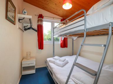 A bedroom with a metal bunk bed a nightstand with a lamp a window with red curtains and an iron on a wall shelf at Newtrain Bay near St Merryn