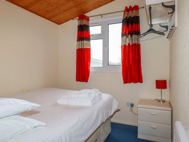 A bedroom with a bed with white linens folded towels on it a window with red and brown curtains a bedside table with a red lamp and a wall-mounted clothes rack at Newtrain Bay near St Merryn