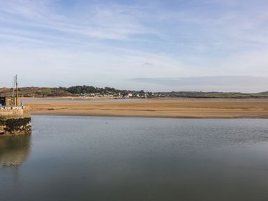 A river with a stone pier and a slow speed sign and a sandy shore with houses in the distance at Constantine Bay near St Merryn