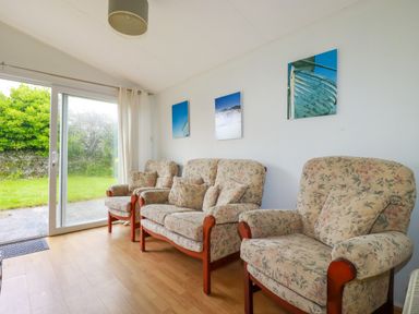 A sitting room with floral armchairs and sofa and sliding glass door opening to a garden at Porthcothan Bay in St Merryn