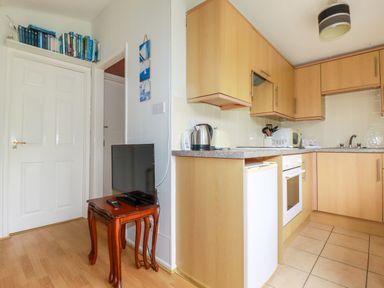 A kitchen with wooden cabinets a countertop with a kettle toaster and microwave a small fridge an oven a small wooden table with a television and books on a shelf above a door at Porthcothan Bay in St Merryn