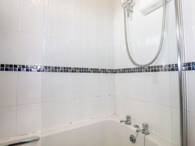 A bathroom shower area with white tiles and a strip of small dark tiles above a bathtub at Porthcothan Bay in St Merryn