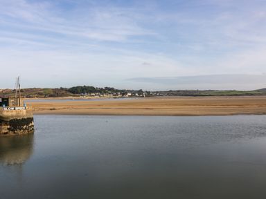 A river with a concrete structure labeled slow speed and a sandy shore with houses and hills in the background at Porthcothan Bay in St Merryn