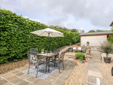 An outdoor patio with a table and chairs under an umbrella surrounded by plants and a wooden shed at Holly Cottage Llechwedd near Conwy