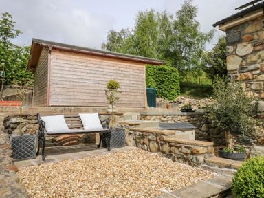 An outdoor seating area with a wooden bench with two white cushions and potted plants on a gravelled surface at Holly Cottage in Llechwedd near Conwy