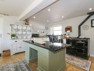 A kitchen with a green island, black stove, white cabinet with plates and mugs, and wooden floor at Holly Cottage in Llechwedd near Conwy