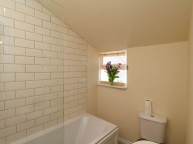 A bathroom with a bathtub, tiled wall, window with blinds and flowers, and a toilet with two rolls of toilet paper on top at Flat 1 in Scarborough