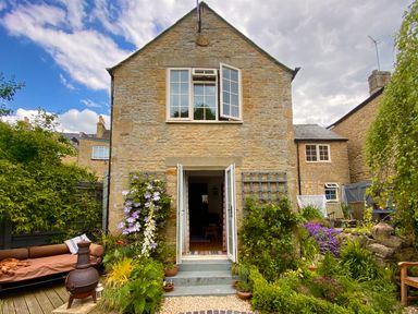 Stone house with open white double doors leading to a garden with plants and a brown outdoor sofa at Lamorna Cottage in Chipping Norton